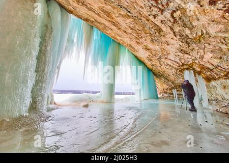 Photographer in ice cave on frozen lake capturing image of large frozen waterfall and blue icicles Stock Photo