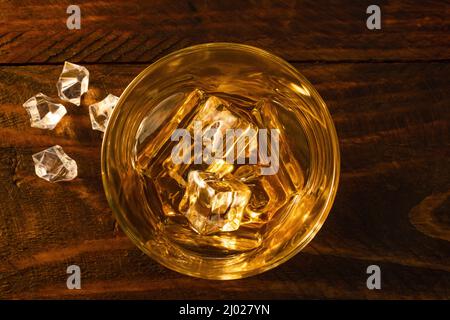 View from above a glass of whiskey on ice with ice cubes lying next to it. Dark wooden background Stock Photo