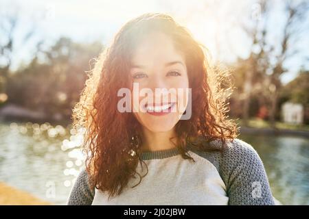 The summer sun is why I love the outdoors. Portrait of a beautiful young woman standing next to a lake. Stock Photo