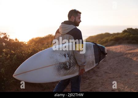 handsome young man in jacket walking at sunset near the beach with his surfboard under his arm, leisure and hobbies concept, copy space for text Stock Photo