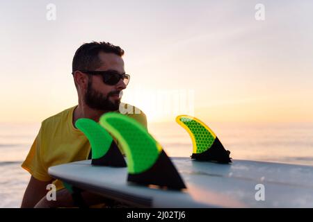 handsome young man in sunglasses sitting by the sea at sunset with his surfboard, leisure and hobbies concept, copy space for text Stock Photo
