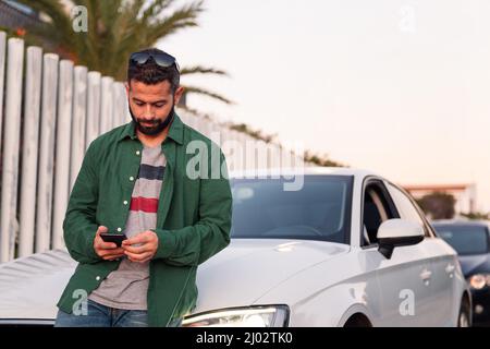 attractive young bearded man sitting on car hood consulting mobile phone, leisure and communication technology concept, copy space for text Stock Photo
