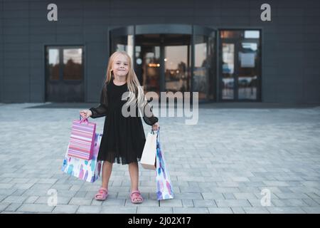 Cute smiling child girl 4-5 year old hold paper bags shopping in store mall making purchases. Buy clothes with season sales. Prepare for birthday part Stock Photo
