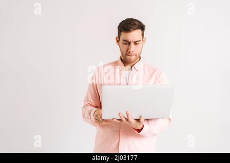 Portrait of serious young man standing with laptop and typing on keyboard, doing freelance job, writing email on white isolated background. Stock Photo