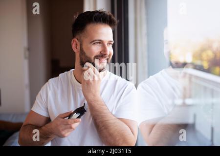 Portrait of a young man shaving his face with an electric razor at home. Stock Photo