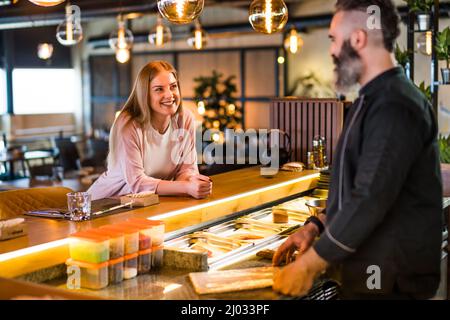 Young woman is making an order at sushi bar. She is talking with sushi chef. Stock Photo