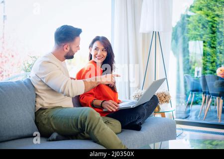 Happy couple relaxing on the couch together. Cheerful woman and handsome man using laptop while browsing on the internet. Handsome man pointing to the Stock Photo