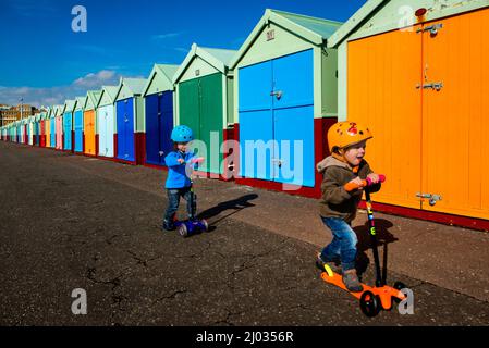 Two boys on their scooters in front of Colourful Beach Huts at Brighton and Hove seafront, Sussex, England, UK Stock Photo