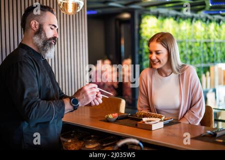 Sushi master is teaching young woman how to use chopsticks at sushi restaurant. Stock Photo