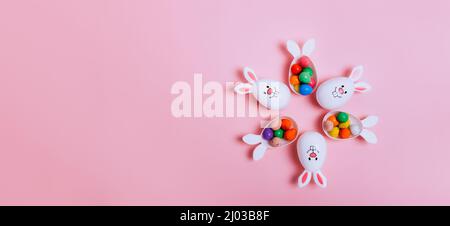 Top view Easter pink background with flower shape made of funny eggs containers with bunny ears filled with candy chocolate eggs. Bunny rabbit symbol. Stock Photo