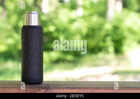 A black thermos with brewed tea or coffee stands on a wooden board with a copyspace. Thermos with hot drink Stock Photo