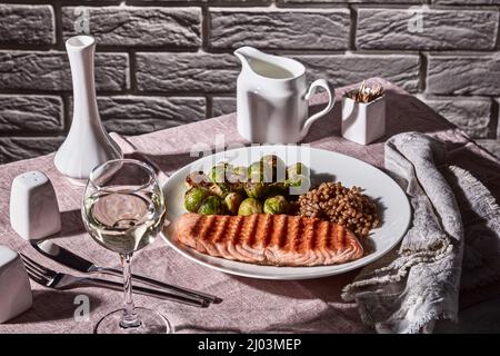 grilled salmon fillet with roast brussel sprouts and cooked farro on a plate with restaurant table setting, white wine in glass, vintage style, brick Stock Photo