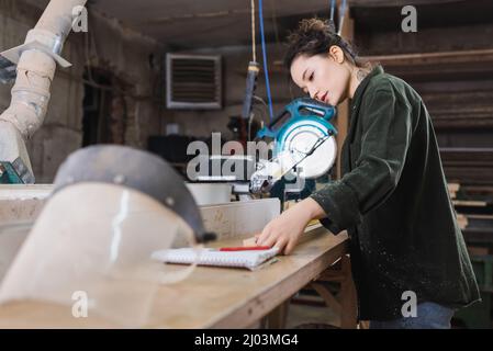 Carpenter measuring wooden plank near blurred safety visor in workshop Stock Photo