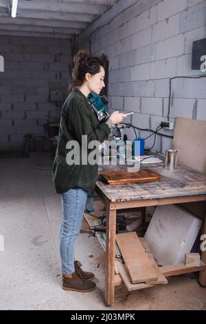 Side view of furniture designer taking photo on cellphone near plank in workshop Stock Photo