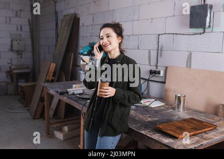 Happy woodworker holding paper cup and talking on cellphone near wooden board in workshop Stock Photo