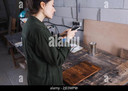 Side view of young craftswoman using smartphone near wooden plank in workshop Stock Photo