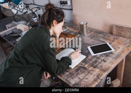 High angle view of carpenter writing on notebook near digital tablet and wood stain in workshop Stock Photo