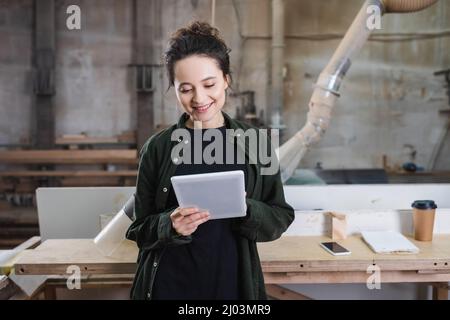 Smiling furniture designer using digital tablet near smartphone and safety visor in workshop Stock Photo