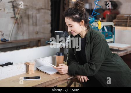 Young carpenter using digital tablet near notebook and coffee to go in workshop Stock Photo