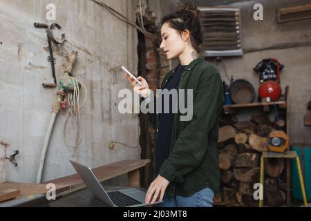 Side view of tattooed woodworker using cellphone near laptop in workshop Stock Photo