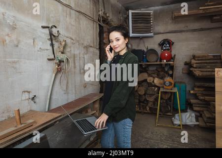 Smiling carpenter looking at camera while talking on smartphone and using laptop in workshop Stock Photo