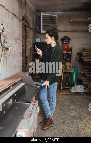 Side view of carpenter using mobile phone near laptop in workshop Stock Photo