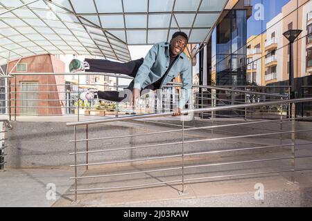 Full length of cheerful young black male with dark hair in stylish clothes smiling while jumping over metal railing near contemporary building on city Stock Photo