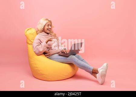 Happy woman relaxing in beanbag chair with laptop, female student using modern gadget, pink background Stock Photo