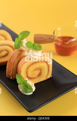 Homemade Pineapple Jam Swiss Roll on Black Ceramic Plate with Cream and Mint on Top. Cake on Yellow Background For Commercial and Ads use for Bakery Stock Photo