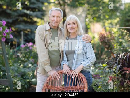 Loving retired spouses embracing, resting in garden in countryside, standing near wicker chair and smiling at camera Stock Photo