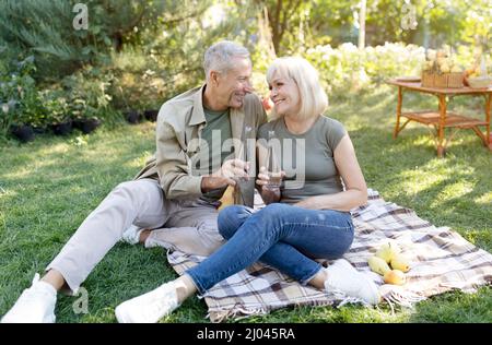 Senior spouses clinking bottles with fresh water, having picnic and sitting on blanket in their garden in countryside Stock Photo
