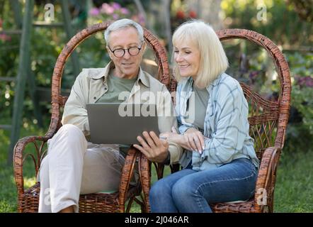 Modern senior spouses relaxing in wicker chairs in open air and using laptop computer, enjoying spring evening outdoors Stock Photo