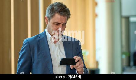 Business portrait - confident businessman standing in office, using phone. Happy mid adult man in shirt and jacket, smiling. Bearded, gray hair. Stock Photo