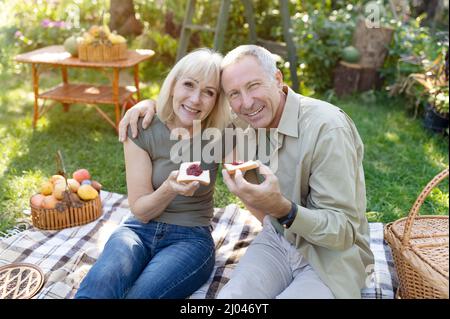 Loving elderly spouses having picnic, sitting on blanket in garden and eating toasts with jam, smiling at camera Stock Photo