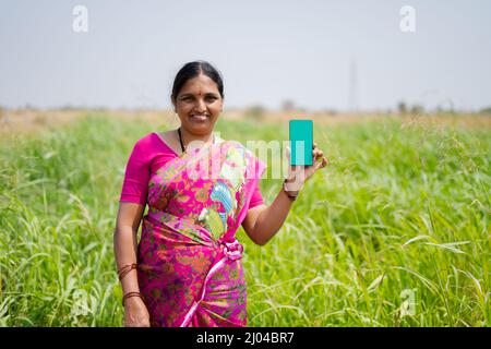 happy smiling Indian woman farmer showing green screen mobile phone in front of green farmland - concept of app advertisement and farm product Stock Photo