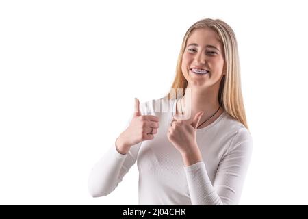 Young pretty girl with braces is showing thumbs up, isolated on white. Stock Photo