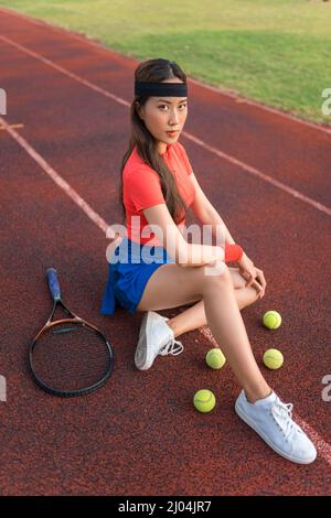 Asian young pretty woman with black headband sitting on the sport stadium with the tennis racket and balls. Sport outdoors. High quality photo Stock Photo