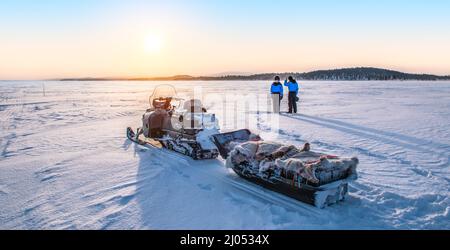 Young couple enjoy sunrise near snowmobile on frozen lake Inari in Finland, Lapland. Stock Photo