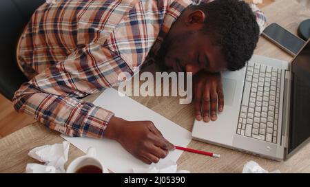 Tired man at home office falling asleep on table with laptop computer, crumpled sheets of paper Stock Photo