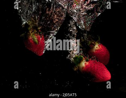 Red strawberries, yellow lemons and blueberries fall into the water. Water splash, drops. Slow movement, black background. Stock Photo