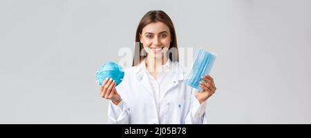 Covid-19, preventing virus, healthcare workers concept. Smiling attractive female doctor in white scrubs showing two variants of personal protective Stock Photo