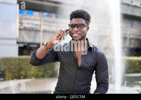 Smiling afro man talking to the mobile outdoors next to a fountain Stock Photo