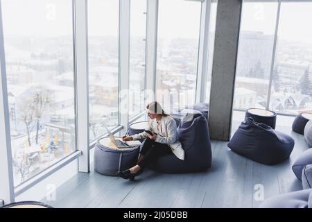 Focused female freelancer sitting in modern workspace with open netbook and checking notifications on mobile phone in casual clothes. Stock Photo
