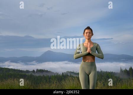 Beautiful young woman meditating with closed eyes with background of green mountains. Peaceful lady standing with namaste gesture and enjoying harmony on nature. Stock Photo