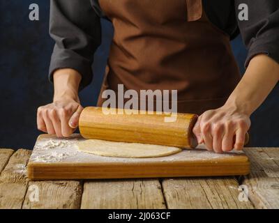 Woman chef in dark uniform rolls out dough on wooden cutting board, dark background. Macro shot. Dough products, recipes for making bread, pizza, past Stock Photo