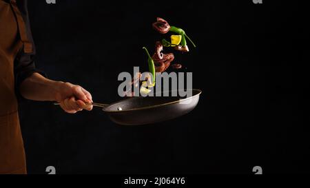 The chef's hand holds a frying pan with seafood and vegetables, lemon slices. Levitation, black background. Healthy food, healthy lifestyle. Food back Stock Photo
