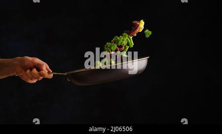 A professional chef cooks octopus with vegetables, herbs and lemon slices in a frying pan. Levitation. Black background. Oriental cuisine, seafood rec Stock Photo