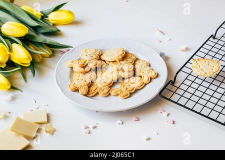 Plate with Easter cookies in bunnies shape next to bunch of yellow tulips on white table. Top view. Easter holidays backing Stock Photo