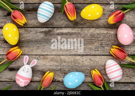 Colorful Easter eggs, one egg with cute bunny face and rabbit ears next to orange-yellow tulips on a rustic wooden table. Easter concept Stock Photo
