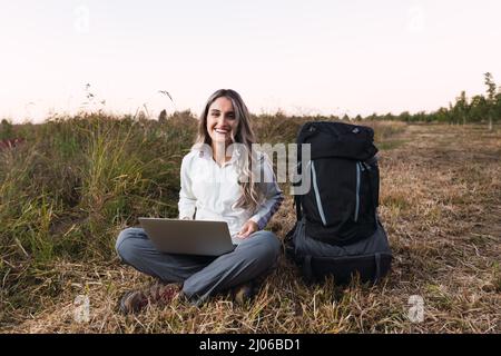 Young woman with a backpack beside her, using a laptop for teleworking in the middle of the field. Digital nomad. Stock Photo